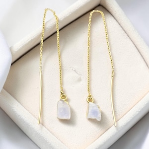 Puede incluir: Un par de pendientes colgantes dorados. Cada pendiente presenta una fina cadena dorada con una pequeña piedra preciosa azul pálido de forma irregular. Los pendientes se muestran en una caja de joyería blanca.