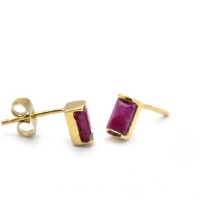 May include: A pair of gold stud earrings featuring rectangular, deep red gemstones. The earrings are set in a gold bezel setting and have a post and butterfly backing. The earrings are displayed on a white background.