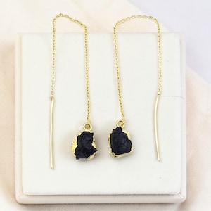 Puede incluir: Un par de pendientes colgantes dorados con colgantes de piedra negra de forma irregular. Los pendientes se muestran sobre una superficie blanca, con una tela suave de color blanquecino en el fondo.
