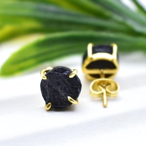 May include: Close-up of a pair of stud earrings. Each earring features a rough, dark gray stone set in a gold-colored metal frame. The earrings are displayed on a white surface with green foliage in the background.
