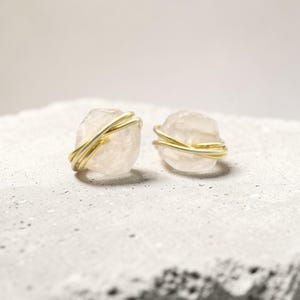 May include: A pair of rose quartz earrings. Each earring features a raw, light pink stone wrapped with gold-colored wire. The earrings are displayed on a textured, light gray surface.