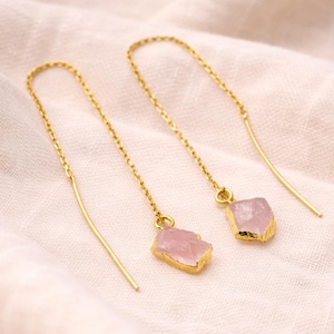 Puede incluir: Un par de pendientes colgantes dorados con piedras de cuarzo rosa en bruto, de color rosa claro. Los pendientes tienen una cadena delicada y una barra curva simple en el extremo. Las piedras son de forma irregular y tienen bordes dorados.