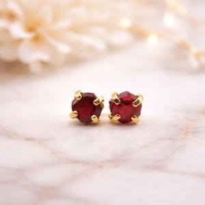 Peut inclure: Une paire de boucles d'oreilles à clous avec des pierres précieuses rouge foncé à facettes serties dans des griffes dorées. Les boucles d'oreilles sont présentées sur une surface en marbre.