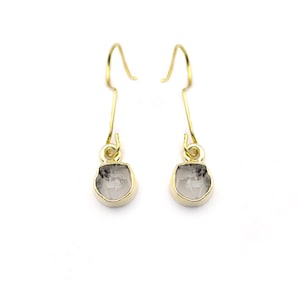 May include: A pair of gold-coloured drop earrings. Each earring features a clear, irregularly shaped stone with grey inclusions, set in a gold-coloured bezel. The earrings hang from gold-coloured ear wires, showcasing a minimalist design.
