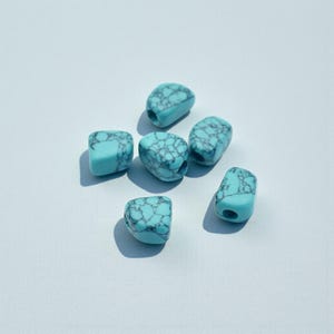 May include: Six turquoise-colored beads with a marbled, dark gray pattern. Each bead is a rounded rectangle shape with a hole drilled through the center. The beads are scattered on a light blue surface.