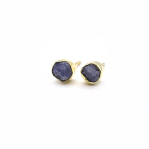 May include: A pair of gold-colored stud earrings, each featuring a rough-cut, blue-purple gemstone. The gemstones are set in a gold bezel, creating a contrast of color and texture. The earrings are displayed on a white background.