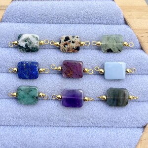 Peut inclure: Neuf perles de pierres pr&eacute;cieuses de diff&eacute;rentes couleurs, dont vert, bleu, violet et gris, sont pr&eacute;sent&eacute;es. Chaque perle rectangulaire est reli&eacute;e par des boucles de fil dor&eacute;. Les perles sont dispos&eacute;es sur une surface textur&eacute;e bleu clair.