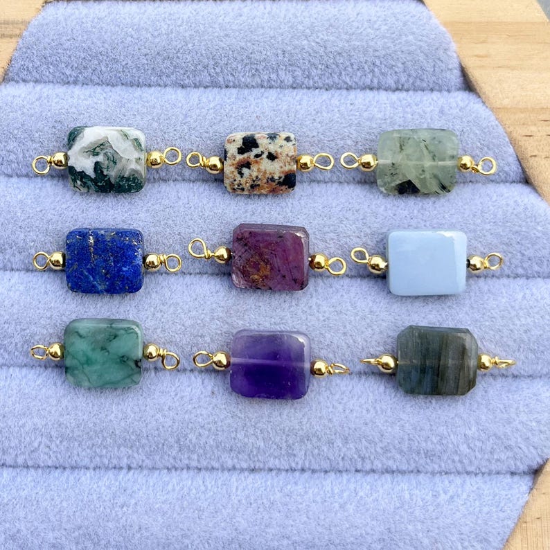 Peut inclure: Neuf perles de pierres pr&eacute;cieuses de diff&eacute;rentes couleurs, dont vert, bleu, violet et gris, sont pr&eacute;sent&eacute;es. Chaque perle rectangulaire est reli&eacute;e par des boucles de fil dor&eacute;. Les perles sont dispos&eacute;es sur une surface textur&eacute;e bleu clair.