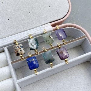 Peut inclure: Une collection de huit pendentifs en pierres pr&eacute;cieuses de diff&eacute;rentes couleurs, dont le marron, le vert, le bleu et le violet. Chaque pierre rectangulaire est reli&eacute;e &agrave; une boucle dor&eacute;e. Les pendentifs sont expos&eacute;s dans une bo&icirc;te &agrave; bijoux rose.