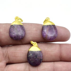 Puede incluir: Tres colgantes de piedras preciosas moradas con tapas y bucles dorados. Los colgantes tienen una forma lisa y redondeada. Las piedras preciosas tienen un aspecto natural y texturizado.
