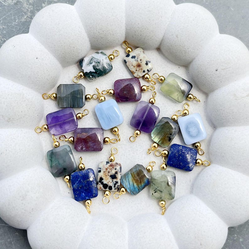 Peut inclure: Une collection de pendentifs en pierres pr&eacute;cieuses de diff&eacute;rentes couleurs, dont le vert, le violet, le bleu et le blanc. Chaque pierre rectangulaire est fix&eacute;e &agrave; une b&eacute;li&egrave;re dor&eacute;e. Les pendentifs sont dispos&eacute;s sur une surface blanche textur&eacute;e.