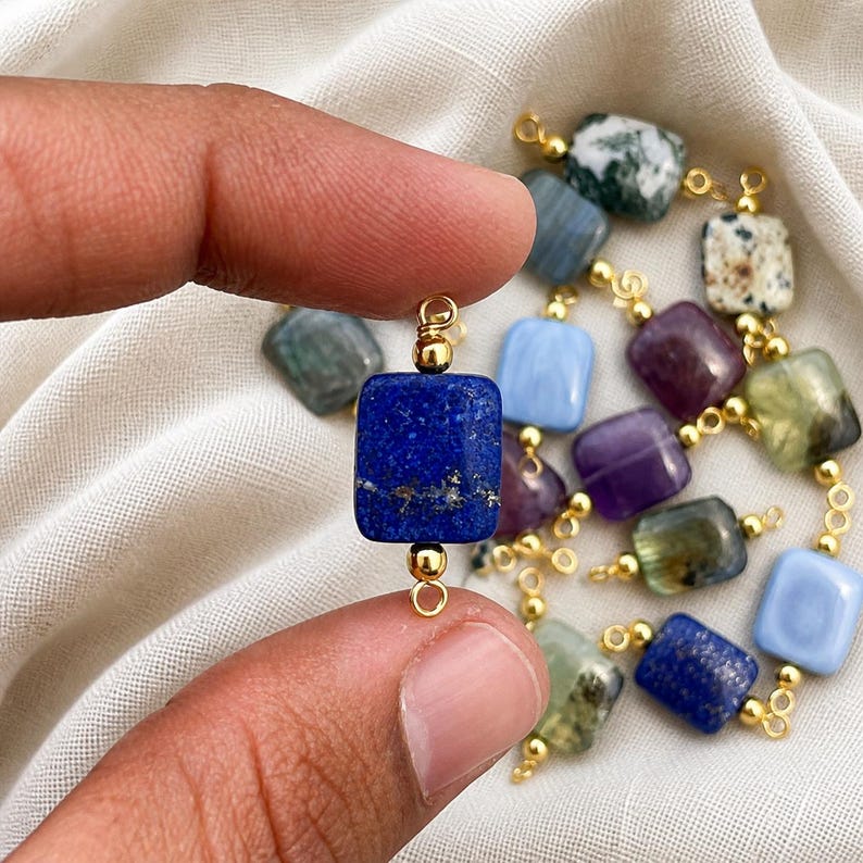Peut inclure: Une collection de perles de pierres pr&eacute;cieuses de diff&eacute;rentes couleurs, dont le bleu, le violet et le vert. Chaque perle est carr&eacute;e ou rectangulaire et reli&eacute;e par des attaches dor&eacute;es. Les perles sont tenues entre les doigts sur un fond de tissu blanc.