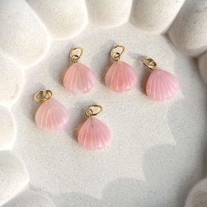 May include: Five pink seashell charms with gold-colored loops. The charms are carved with a ribbed texture, and the gold loops are attached at the top. The charms are arranged on a white surface.