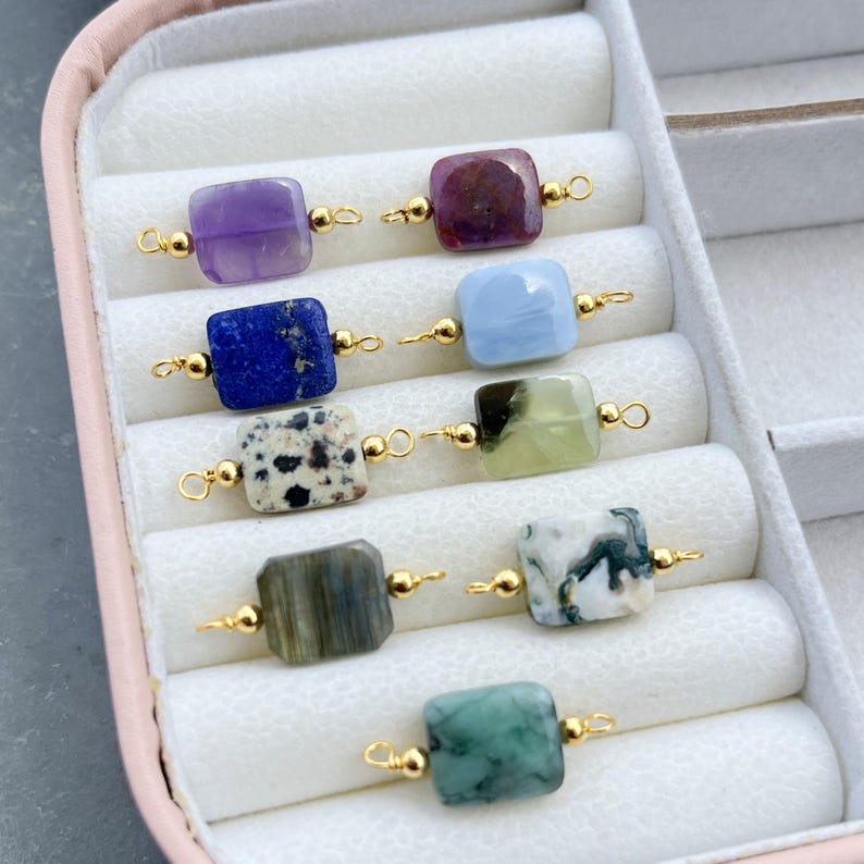Peut inclure: Collection de perles carr&eacute;es en pierres pr&eacute;cieuses de diff&eacute;rentes couleurs, notamment violet, bleu et vert, reli&eacute;es par des attaches dor&eacute;es. Les perles sont pr&eacute;sent&eacute;es dans une bo&icirc;te &agrave; bijoux rose.
