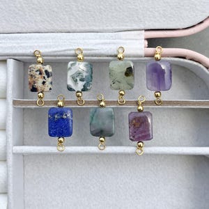 Peut inclure: Une collection de huit pendentifs en pierres pr&eacute;cieuses de diff&eacute;rentes couleurs et motifs. Chaque pierre rectangulaire est attach&eacute;e &agrave; une boucle en m&eacute;tal dor&eacute;. Les pierres pr&eacute;cieuses comprennent des vari&eacute;t&eacute;s marron mouchet&eacute;, blanc et vert, bleu et violet. Les pendentifs sont expos&eacute;s dans un &eacute;crin blanc.