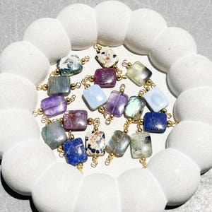 Peut inclure: Collection de pierres pr&eacute;cieuses carr&eacute;es de diff&eacute;rentes couleurs, notamment violet, bleu, vert et blanc, avec des boucles en m&eacute;tal dor&eacute;. Les pierres pr&eacute;cieuses sont dispos&eacute;es sur un plateau blanc en forme de nuage. Id&eacute;al pour la cr&eacute;ation de bijoux.