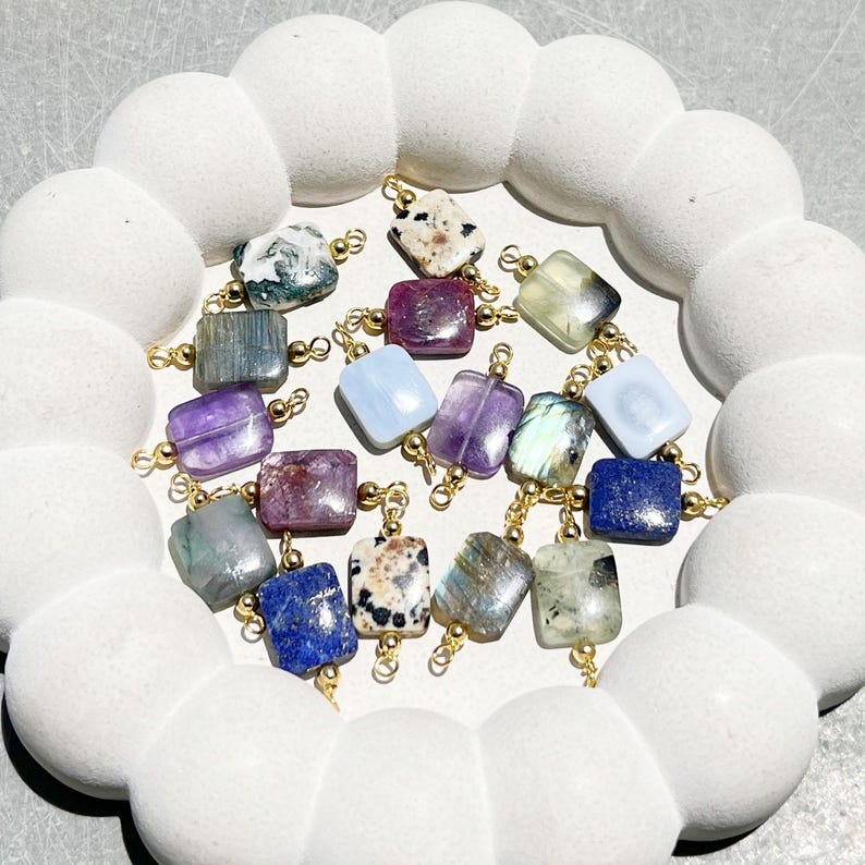 Peut inclure: Collection de pierres pr&eacute;cieuses carr&eacute;es de diff&eacute;rentes couleurs, notamment violet, bleu, vert et blanc, avec des boucles en m&eacute;tal dor&eacute;. Les pierres pr&eacute;cieuses sont dispos&eacute;es sur un plateau blanc en forme de nuage. Id&eacute;al pour la cr&eacute;ation de bijoux.