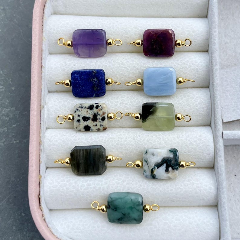 Peut inclure: Une s&eacute;lection de perles de pierres pr&eacute;cieuses de diff&eacute;rentes couleurs, dont le violet, le bleu, le vert et des perles tachet&eacute;es. Chaque perle rectangulaire est reli&eacute;e par des boucles de fil dor&eacute;, composants de fabrication de bijoux.