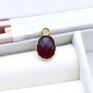Könnte beinhalten: Ein facettierter, oval geformter, dunkelroter Edelsteinanhänger mit goldfarbener Lünette und kleiner Schlaufe oben. Der Anhänger ist vor einem weißen Hintergrund platziert, der seine satte Farbe und den aufwendigen Schliff hervorhebt. Dieses Schmuckstück eignet sich für Halsketten oder Ohrringe.