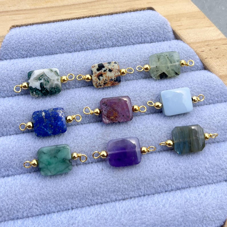 Peut inclure: Neuf perles carr&eacute;es en pierres pr&eacute;cieuses de diff&eacute;rentes couleurs, dont vert, bleu, violet et marron mouchet&eacute;, sont reli&eacute;es par de petites boucles dor&eacute;es. Les perles sont dispos&eacute;es en trois rang&eacute;es de trois, reposant sur une surface bleu clair.