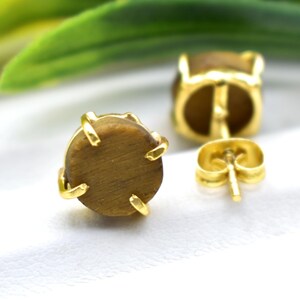May include: Close-up of a pair of gold-colored stud earrings. Each earring features a round, brown gemstone held in place by four gold prongs. The earrings are set against a white background, with a blurred green leaf in the background.