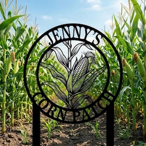 May include: Black metal sign reading "JENNY'S CORN PATCH" in a circular design. The centre shows an illustration of an ear of corn with leaves. The sign is set in a cornfield under a blue sky.