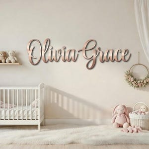 Könnte beinhalten: Eine Szene im Kinderzimmer mit einem weißen Kinderbett, Plüschtieren und einer roségoldenen Wanddekoration mit der Aufschrift "Olivia Grace". Ein Regal beherbergt Teddybären. Ein Blumenkranz und ein weißer Teppich vervollständigen die Einrichtung.