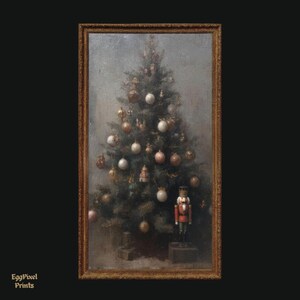 Op de afbeelding: Een ingelijst vintage schilderij van een versierde kerstboom. De boom is versierd met ornamenten in tinten goud, wit en roze. Een rood-witte notenkraker staat aan de voet van de boom. De lijst is goudkleurig en de achtergrond is grijs.