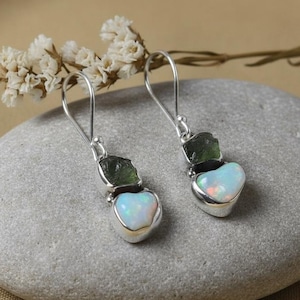 Puede incluir: Pendientes colgantes de plata con un ópalo en forma de corazón y una piedra verde. Los pendientes se exhiben sobre una piedra gris con un fondo neutro. Pequeñas flores blancas en la esquina superior izquierda.
