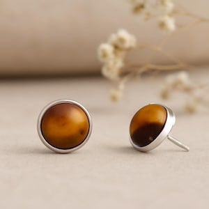 Pode incluir: Um par de brincos de pressão com uma pedra redonda de cor âmbar embutida em uma moldura prateada. Os brincos têm um pino e um fecho para uso seguro. Os tons quentes das pedras contrastam com o metal frio.