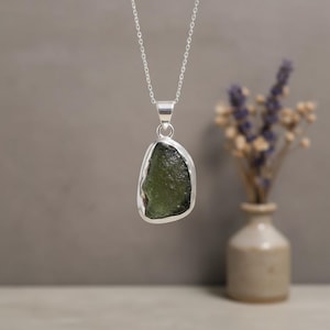 May include: A silver pendant necklace featuring a raw, olive-green moldavite stone. The stone is irregularly shaped and set in a polished silver bezel. The necklace hangs against a neutral background, with a blurred vase of dried flowers in the background.