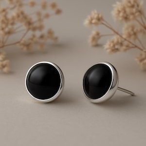 Puede incluir: Un par de pendientes de botón con una piedra negra pulida engastada en metal plateado. Los pendientes tienen un diseño sencillo y moderno con forma redondeada. El fondo es de color neutro con suaves acentos florales borrosos.