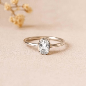 Puede incluir: Un anillo de plata con una gema ovalada y transparente. El anillo está sobre una superficie rosa claro, con un elemento floral borroso en el fondo. La gema es el punto focal, mostrando su claridad y forma. La banda del anillo es delgada y pulida.