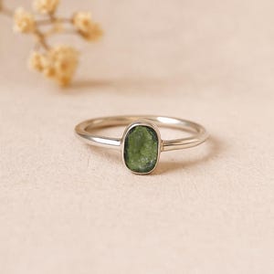 Peut inclure: Une bague en argent avec une pierre verte ovale. La bague a un anneau fin et un design simple. La pierre verte est sertie dans une lunette minimaliste. La bague est probablement en argent sterling. Le design est élégant et discret.