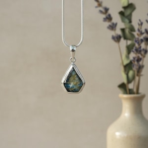 Puede incluir: Un collar con colgante de plata con un colgante en forma de lágrima. El colgante tiene un marco plateado y un centro azul y dorado. El collar se muestra frente a un jarrón con ramitas de lavanda.