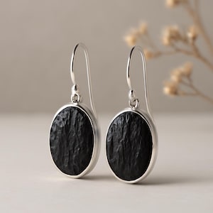 May include: A pair of silver drop earrings featuring oval, textured black stones. The earrings have a simple, elegant design with a hook closure. The black stones have a rough, organic appearance, set in a polished silver frame.