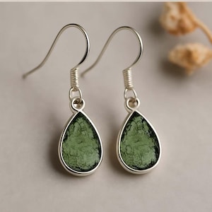 Puede incluir: Pendientes colgantes de plata con piedras verdes en forma de lágrima. Las piedras tienen un aspecto texturizado y están engastadas en un bisel de plata. Los pendientes tienen un diseño de gancho simple para orejas perforadas.