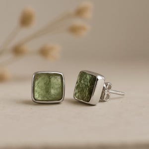 Puede incluir: Un par de pendientes de botón cuadrados con una piedra preciosa verde claro engastada en metal plateado. Los pendientes se muestran sobre una superficie neutra, mostrando su diseño simple pero elegante.