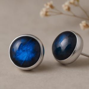 Könnte beinhalten: Ein Paar Ohrstecker mit einem runden, polierten, blauen Edelstein, der in einer silbernen Lünette gefasst ist. Die Edelsteine haben ein wirbelndes, irisierendes Muster. Die Ohrringe werden auf einem neutralen Hintergrund präsentiert.