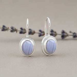 Könnte beinhalten: Ein Paar silberne Ohrhänger mit ovalen, hellblauen Edelsteinen. Die Ohrringe haben ein schlichtes, elegantes Design mit poliertem Finish. Der Hintergrund zeigt eine verschwommene Anordnung von lila Blumen.