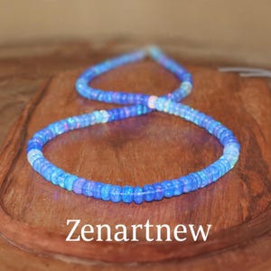 May include: A blue beaded necklace with iridescent highlights. The necklace is composed of small, round beads and is presented on a wooden surface. The text "Zenartnew" is visible at the bottom of the image.