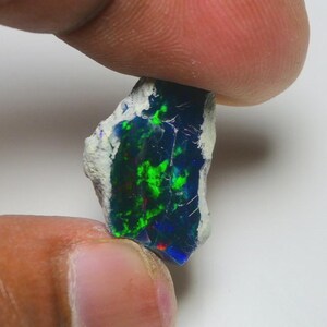 May include: A rough-cut black opal gemstone with vibrant green, blue, and red flashes. The opal is held between fingers, showcasing its natural, unpolished texture and color play. The stone has a white matrix.