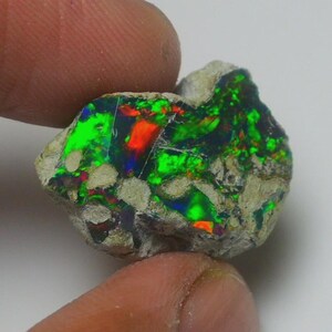May include: A rough-cut black opal gemstone with vibrant flashes of green, red, and blue. The opal is set in a matrix of light gray rock. The gemstone is held between two fingers.
