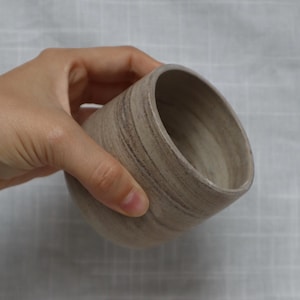 Könnte beinhalten: Handgefertigte, beige Keramiktasse mit abgerundetem Boden und abgeschrägtem Rand. Die Tasse hat eine strukturierte Oberfläche mit dezenten braunen Streifen, gehalten von einer Hand. Perfekt für Kaffee oder Tee.