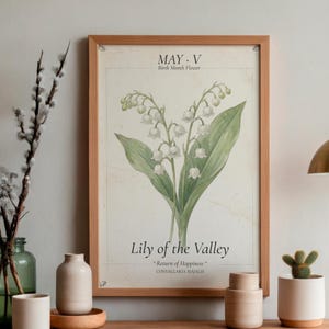 以下が含まれることがあります： スズランのイラストが描かれた額入りの植物画。テキストには「MAY. V Birth Month Flower」と「Lily of the Valley - Return of Happiness - CONVALLARIA MAJALIS」が含まれています。フレームは明るい茶色の木製です。