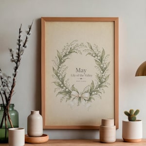 以下が含まれることがあります： スズランの花輪が描かれた額入りのプリント。プリントは明るい茶色の木製フレームで、「May Lily of the Valley」の文字が入っています。木製の表面に飾られており、花瓶と小さなサボテンが置かれています。