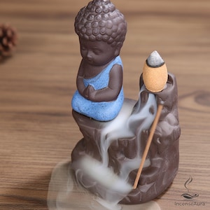 May include: A dark brown ceramic incense burner featuring a small Buddha figure with a blue robe sitting on a rock formation. A lit tan incense cone sits on the top right, with white smoke cascading down the rock surface like a waterfall.