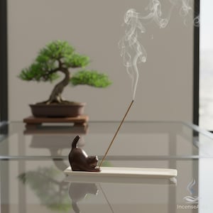 以下が含まれることがあります： 白の長方形のベースに、茶色のセラミック製の猫の香立て。お香が焚かれており、白い煙が出ています。背景には盆栽があります。画像はガラスの表面に配置されています。