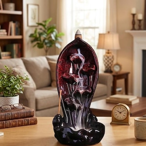 May include: A dark red and black ceramic incense burner shaped like a leaf with a central channel. A lit incense cone sits at the top, producing a white smoke trail that flows downward over sculpted petal-like tiers into a basin at the base.