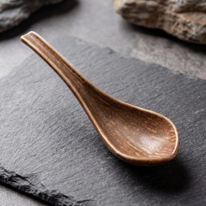 Può includere: Un cucchiaio in ceramica marrone con un lungo manico e una ciotola arrotondata. Il cucchiaio ha una superficie strutturata ed è posto su uno sfondo grigio scuro. Il cucchiaio è progettato per servire zuppe e altri piatti.